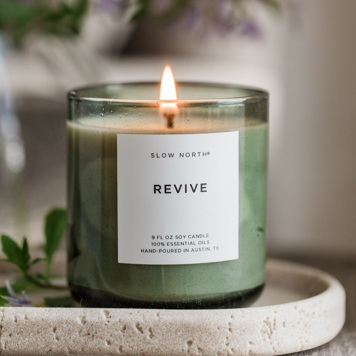 Candle labeled 'Revive' by Slow North on a stone surface with a blurred floral background
