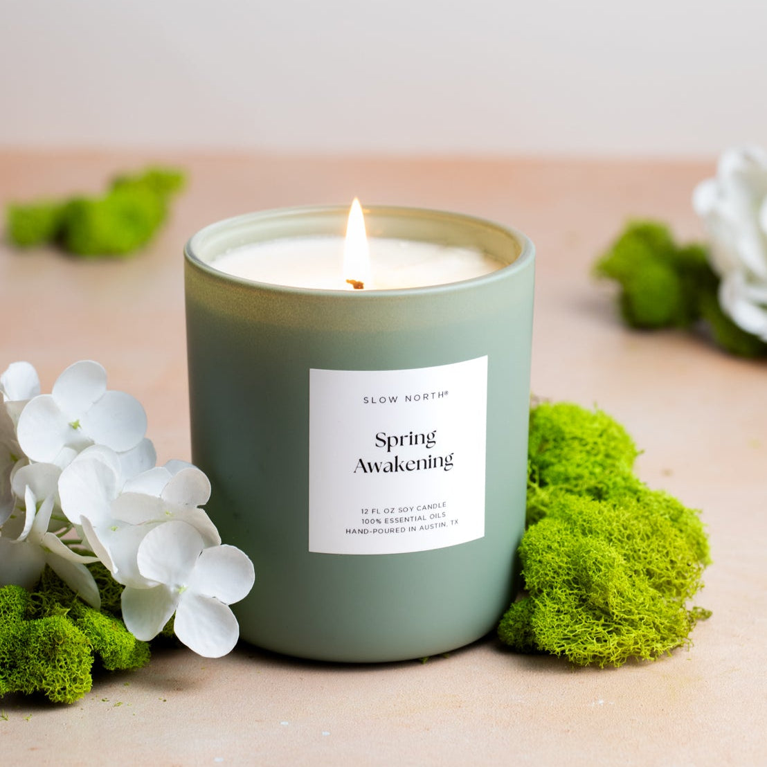 Green candle labeled 'Spring Awakening' surrounded by flowers and moss on a wooden surface.