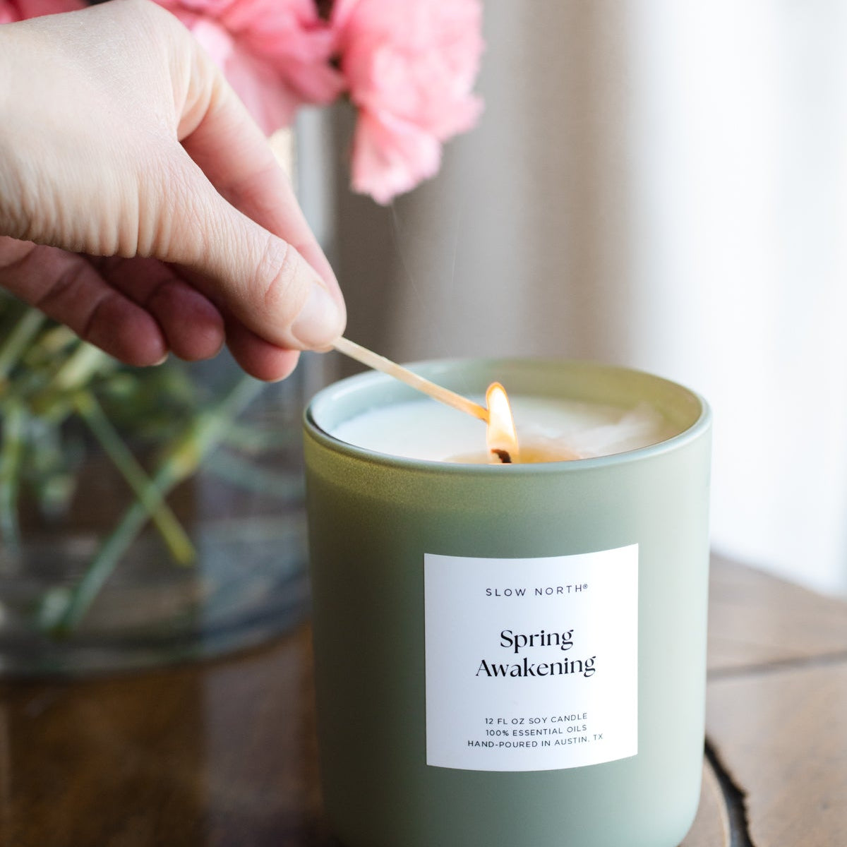 Hand lighting a candle labeled 'Spring Awakening' with pink flowers in the background