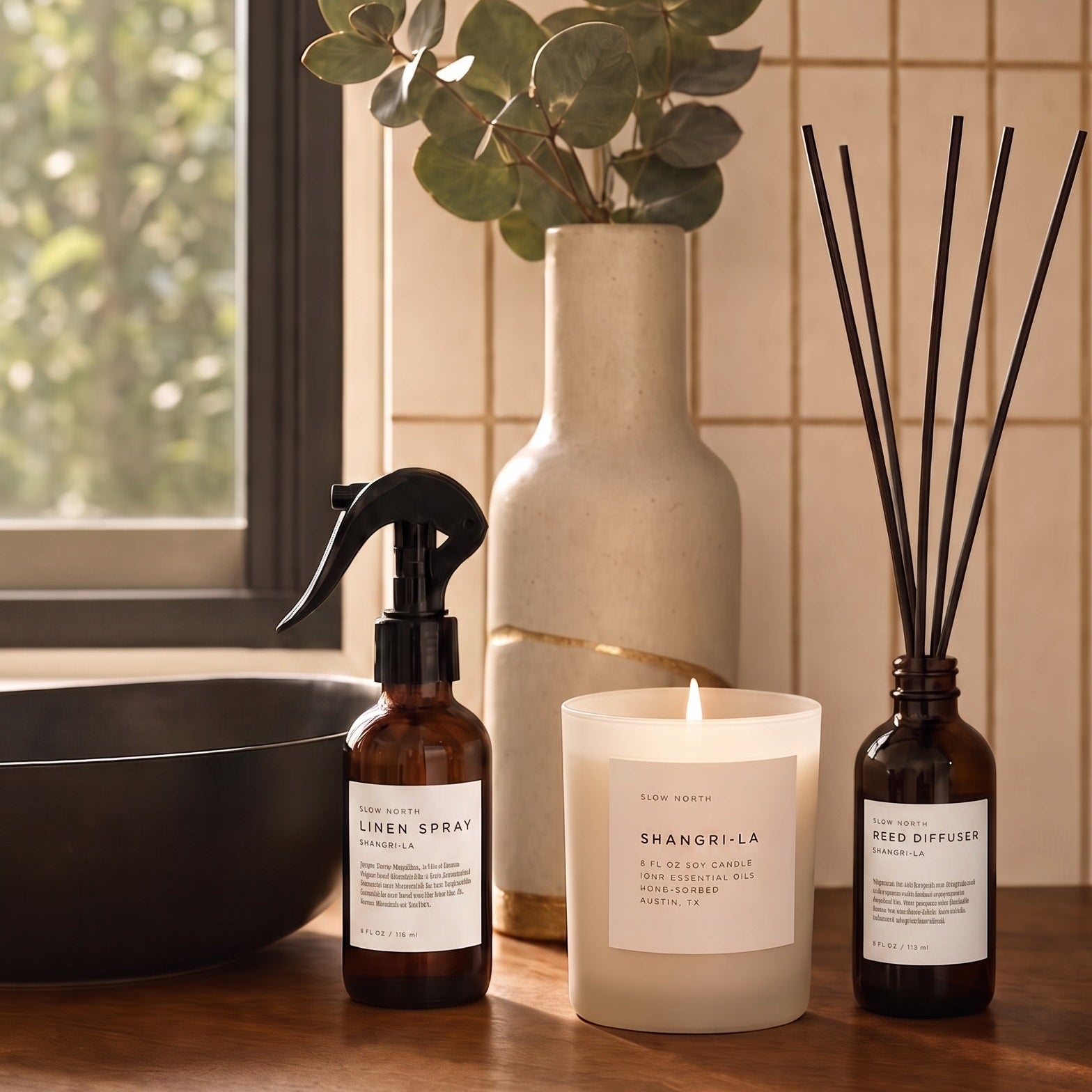 A styled home interior scene on a wooden counter with a lit candle, an amber spray bottle, and an amber reed diffuser bottle, alongside a tall vase with eucalyptus leaves and a window in the background.