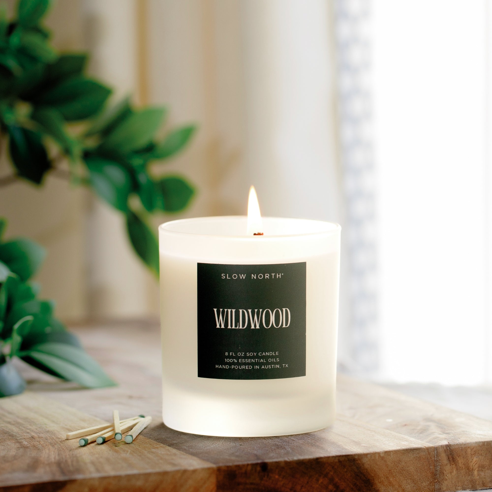 Candle labeled 'Wildwood' by 'Slow North' on a wooden surface with greenery in the background