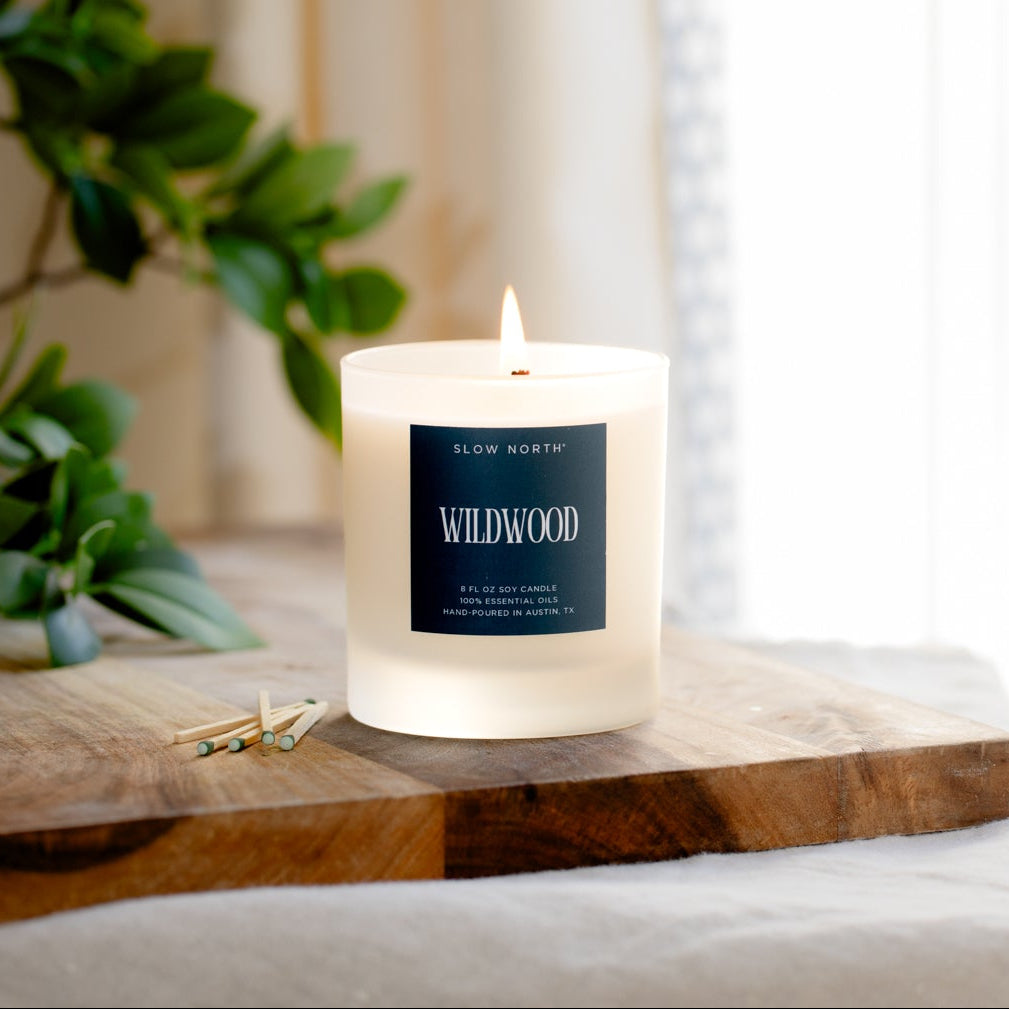 Candle with a 'Wildwood' label on a wooden surface near greenery