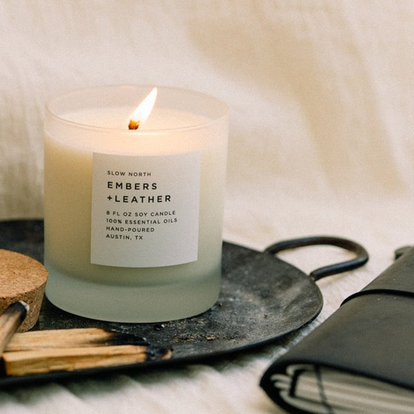 lit off white candle next to cork lid and palo santo on black tray in front of a white sheet background.