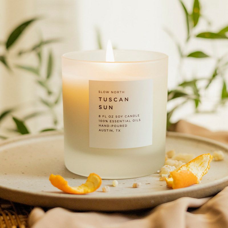Tuscan Sun by Slow North. Lit white candle in frosted glass tumbler on a tan circle tray surrounded by orange peels and greenery.