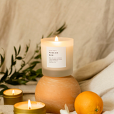 Tuscan Sun by Slow North. Lit white candle in frosted glass tumbler on an orange stand. Background is a tan sheet with green plant and oranges and lit gold travel tin and mini-candle.