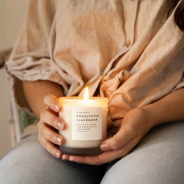 Woman's hands holding lit soy wax candle in 8 ounce clear frosted tumbler on lap. By Slow North