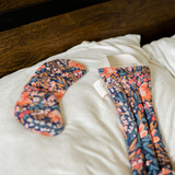 One eye mask and one neck wrap featuring a floral pattern with a dark blue background on a bed with white sheets.
