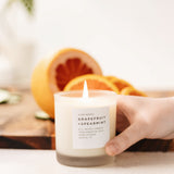 Hand holding lit soy wax candle in 8 ounce clear frosted tumbler by a cutting board with sliced grapefruit. By Slow North