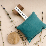A Slow North Lavender + Cedar teal, rectangle sachet looped onto a round wooden block on a tan background. Surrounded by scattered loose lavender buds.