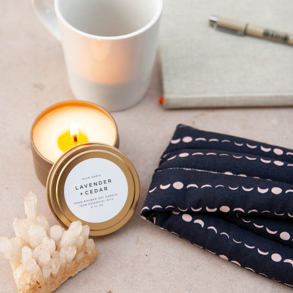 Lavender + Cedar soy wax candle in 6 ounce gold travel tin on desk surrounded by a journal, a white mug, and a solstice print neck wrap. By Slow North