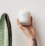 Hand holding lit soy wax candle in 8 ounce clear frosted tumbler near a cactus. By Slow North