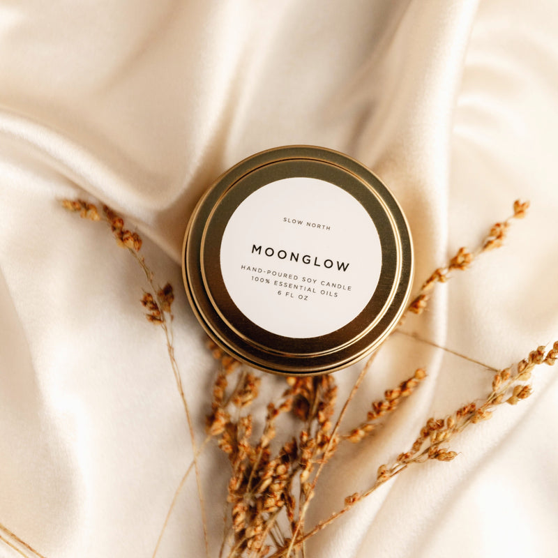 Moonglow soy wax candle in 6 ounce gold travel tin on white silk fabric. Brown floral by base of candle. By Slow North