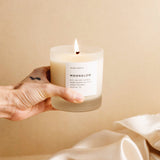 Hand holding lit soy wax candle in 8 ounce clear frosted tumbler over silk fabric and brown floral stems. By Slow North