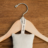 A Slow North cedar sachet hanging on a tan wooden hanger laying flat on a brown wood background