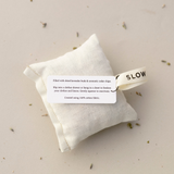 A Slow North rectangle, off-white lavender + cedar sachet laying flat on a tan background. Loose scattered herbs surround the product. The tag is visible. TEXT: Slip into a clothes drawer or hang in a closet to freshen your clothes and linens for insect-repelling benefits. Gently squeeze to reactive.