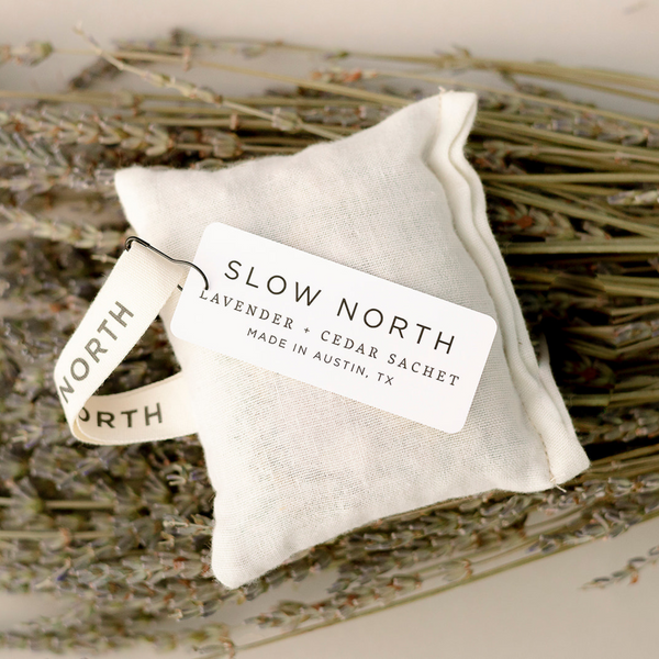A Slow North lavender + cedar rectangle off-white cotton sachet laying flat on a bundle of lavender herbs.