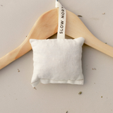 A Slow North lavender + cedar sachet hanging on a tan wooden hanger laying flat on a tan background with scattered loose lavender