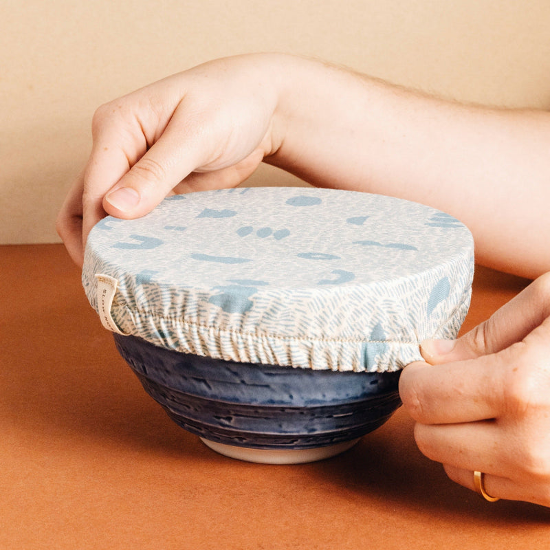 Hands stretching blue and white reusable fabric dish cover over blue bowl by Slow North